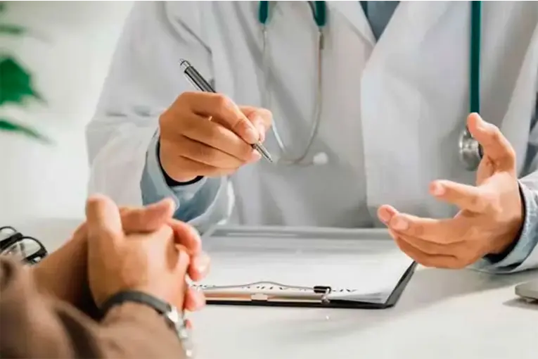 Un médico explica algo a un paciente en un consultorio, con una carpeta y una lapicera sobre el escritorio